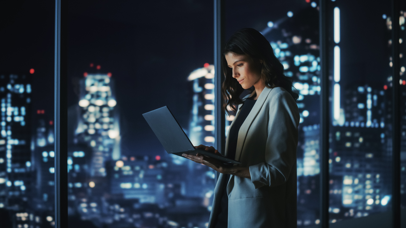 Big City Modern Office at Night: Successful Young Businesswoman Standing and Using Laptop. Beautiful Female Digital Entrepreneur Thinking of Investment Strategy for e Commerce Project.