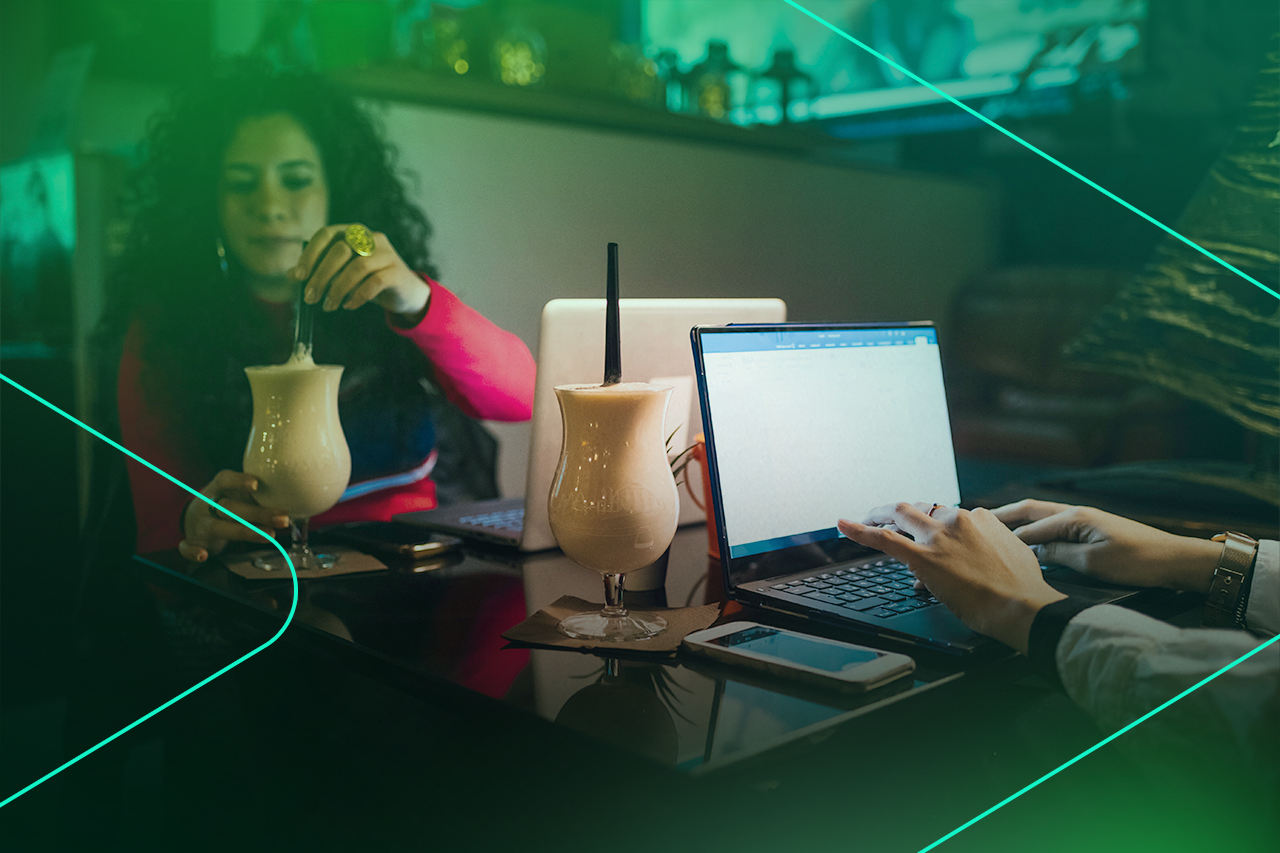 Duas pessoas trabalhando em laptops com bebidas ao fundo.